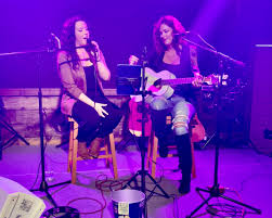 Two women perform on stage under purple lighting; one sings into a microphone while the other plays an acoustic guitar. Both are seated on stools and surrounded by musical equipment.