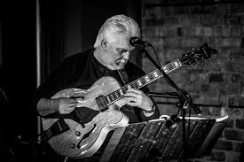 A man with white hair plays an electric guitar on stage, reading music from a stand. The scene is in black and white, with a microphone positioned in front of him and a brick wall in the background.