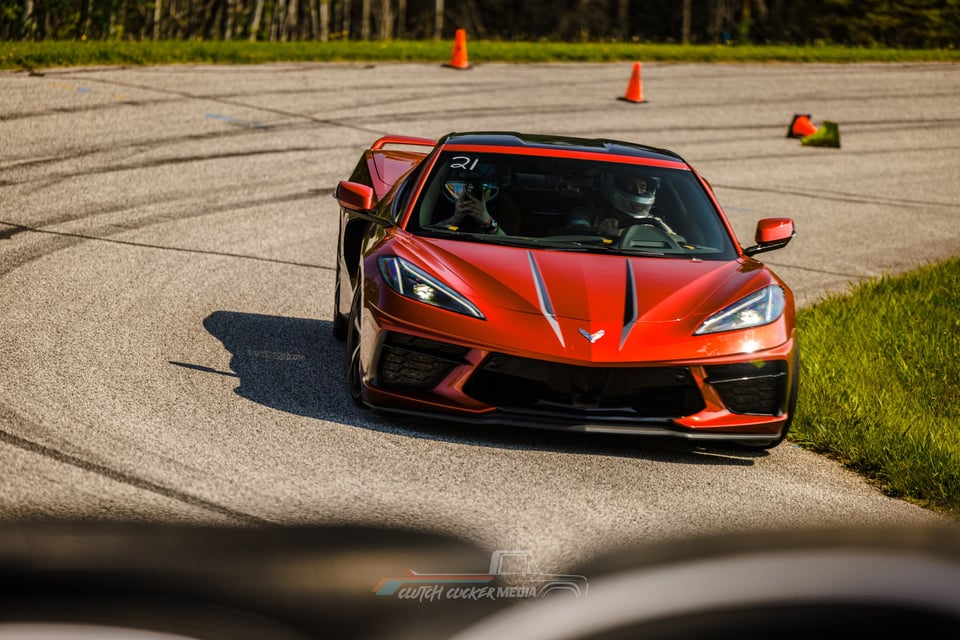 A red Chevrolet Corvette C8 with black racing stripes takes a sharp corner on a racetrack, driven by two people in helmets, with orange cones marking the course.