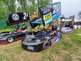 Three small racing go-karts are parked on grass, with large number signs on top. The closest kart displays "70" and a "FOR SALE" sign. People stand in the background near trees.