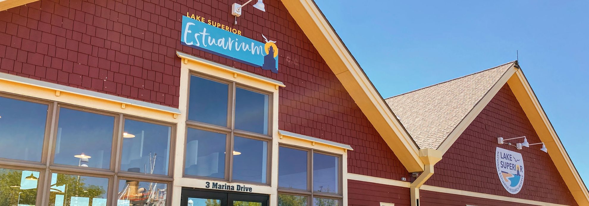A red building with cream trim, large windows, and signs reading "Lake Superior Estuarium" and "Harbor Superior" under a sunny blue sky. The address "3 Marina Drive" is above the entrance.