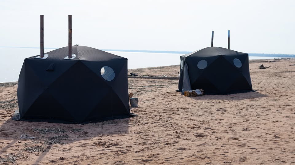 Two black tent-like structures with round windows and metal chimneys stand on a sandy beach near the water, with firewood and buckets nearby under a bright sky.