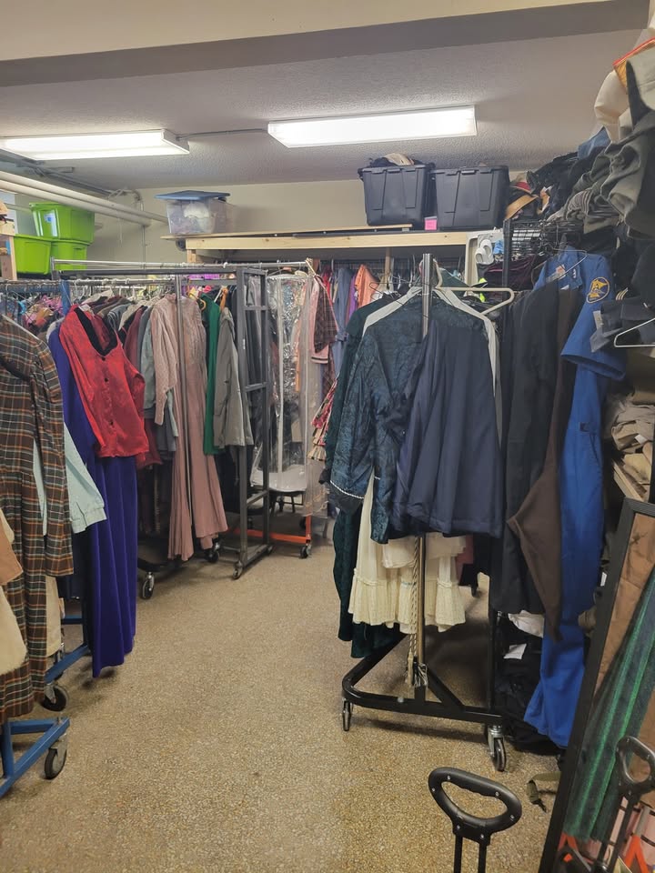 Rows of clothing racks filled with various costumes, including dresses, jackets, and uniforms, are organized in a storage room with fluorescent lighting and plastic bins on shelves above.