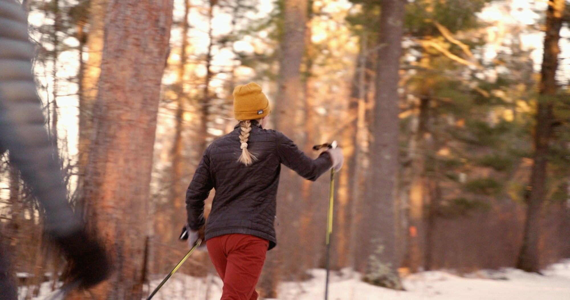 A person with a blond braid, yellow beanie, dark jacket, and red pants is skiing on a snowy path through a forest of tall trees during the Lake Superior Ice Festival, with another skier visible at the edge of the frame.