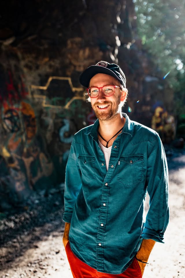 A smiling man wearing glasses, a cap, and a teal button-up shirt stands outdoors in sunlight, with colorful graffiti on a rock wall in the background.