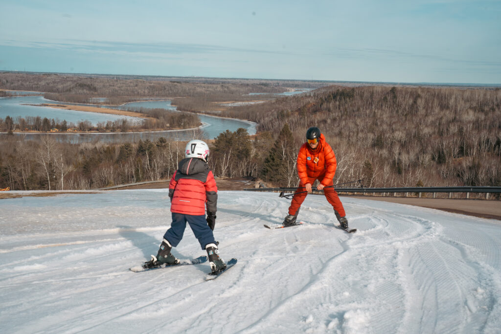 A child and an adult in red winter gear ski down a snowy slope, overlooking a scenic landscape with winding rivers, forest, and distant hills under a blue sky at the Lake Superior Ice Festival.