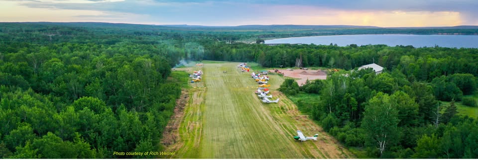 Aerial view of a small grass airstrip surrounded by dense green forest, with several small airplanes parked along the runway near a lake, under a partly cloudy sky at sunset.