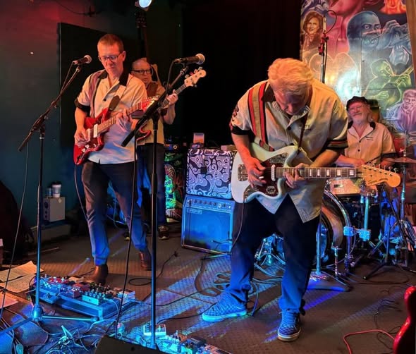 Four older men in casual shirts play electric guitar, bass, and drums on a small, colorful stage decorated with vibrant murals. They are focused on their instruments, performing live music together.