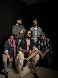 Five men pose together against a dark background; one is seated on a furry chair wearing sunglasses and an open shirt, while the others stand or kneel around him, all dressed in casual, eclectic clothing and accessories.