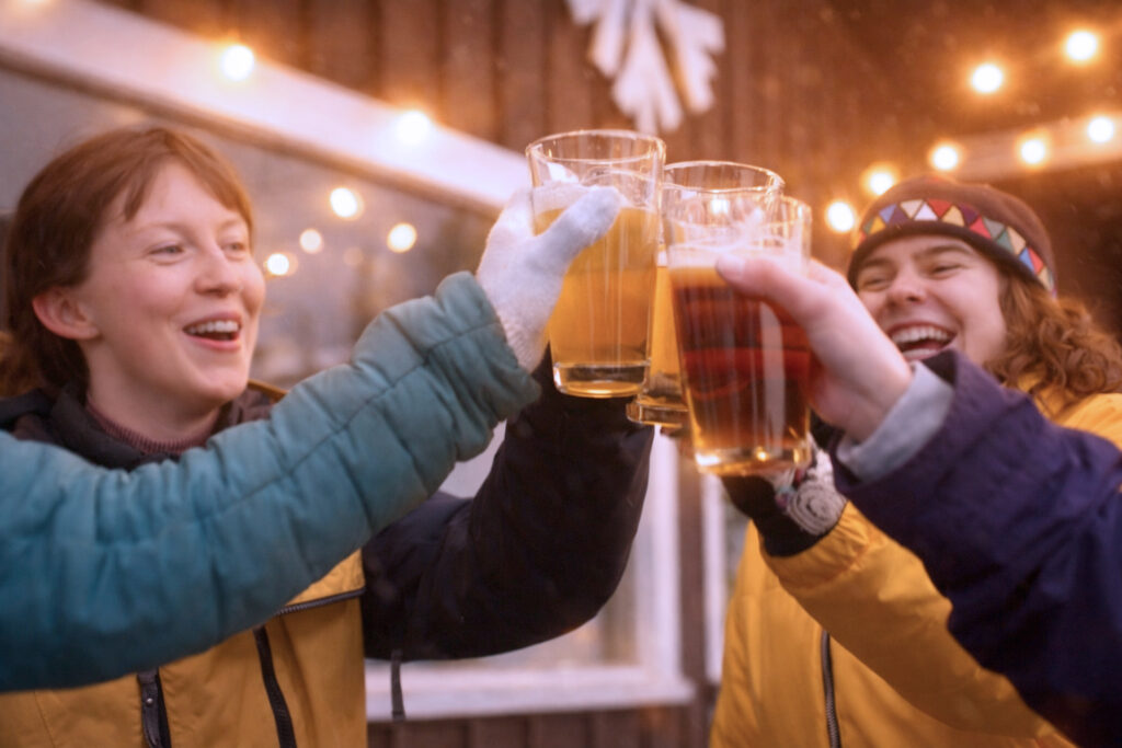 Three people wearing winter jackets and gloves cheerfully clink glasses of beer together outside at the Lake Superior Ice Festival, with warm string lights and a snowflake decoration in the background.