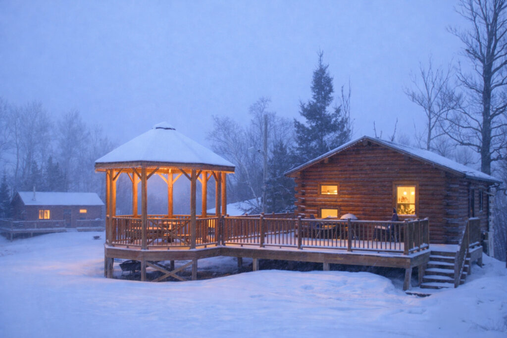 A cozy log cabin and warmly lit wooden gazebo sit surrounded by snow and bare trees on a winter evening, capturing the peaceful, serene charm of a Lake Superior Ice Festival night.