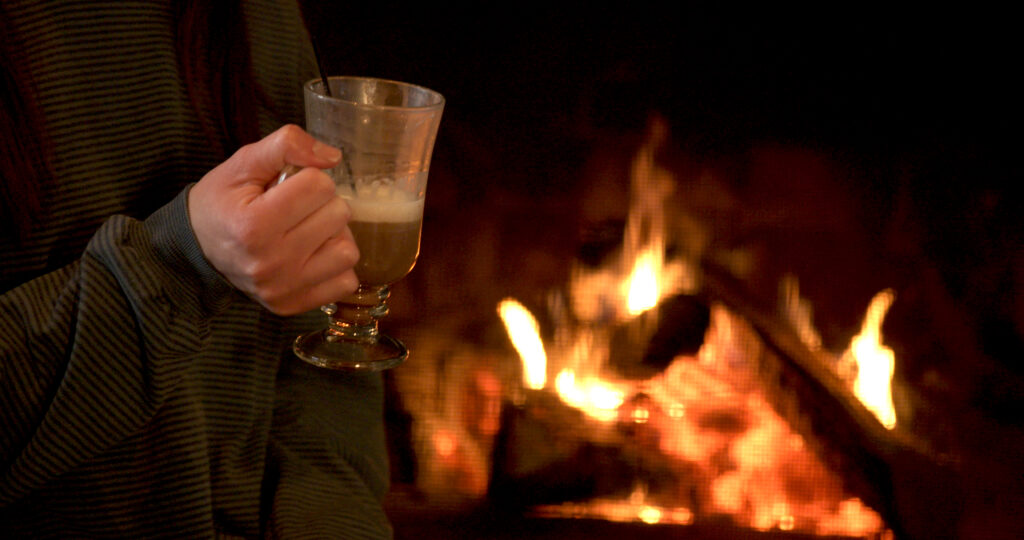 A person in a striped sweater holds a glass mug with a creamy drink, sitting near a warm, glowing fireplace—perfect for unwinding after attending the Lake Superior Ice Festival.