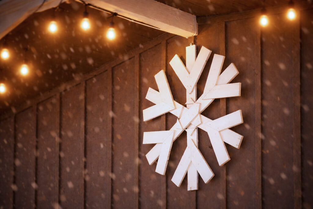 A large white wooden snowflake decoration hangs on a brown wall under warm string lights, with light snow falling in the scene—evoking the cozy spirit of the Lake Superior Ice Festival.