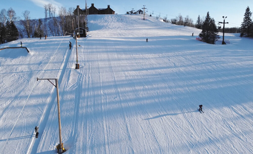 Skiers glide down a snowy ski slope under a clear sky, with ski lifts on the left and a lodge at the top of the hill—capturing the lively spirit of winter like the Lake Superior Ice Festival. Shadows from trees and poles stretch across the snow.