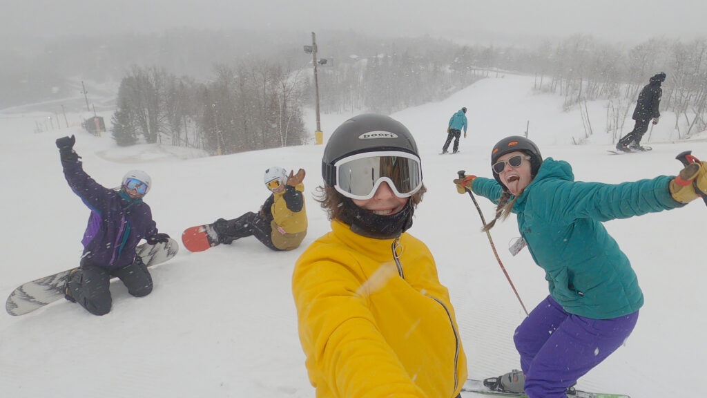 A group of five people in winter gear pose happily on a snowy ski slope at the Lake Superior Ice Festival, with some sitting on snowboards and others standing on skis, surrounded by snow-covered trees.