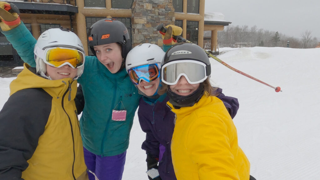 Four people in ski gear and helmets smile and pose together outside a building on a snowy day, with ski goggles and winter jackets, enjoying the Lake Superior Ice Festival at a scenic ski resort.