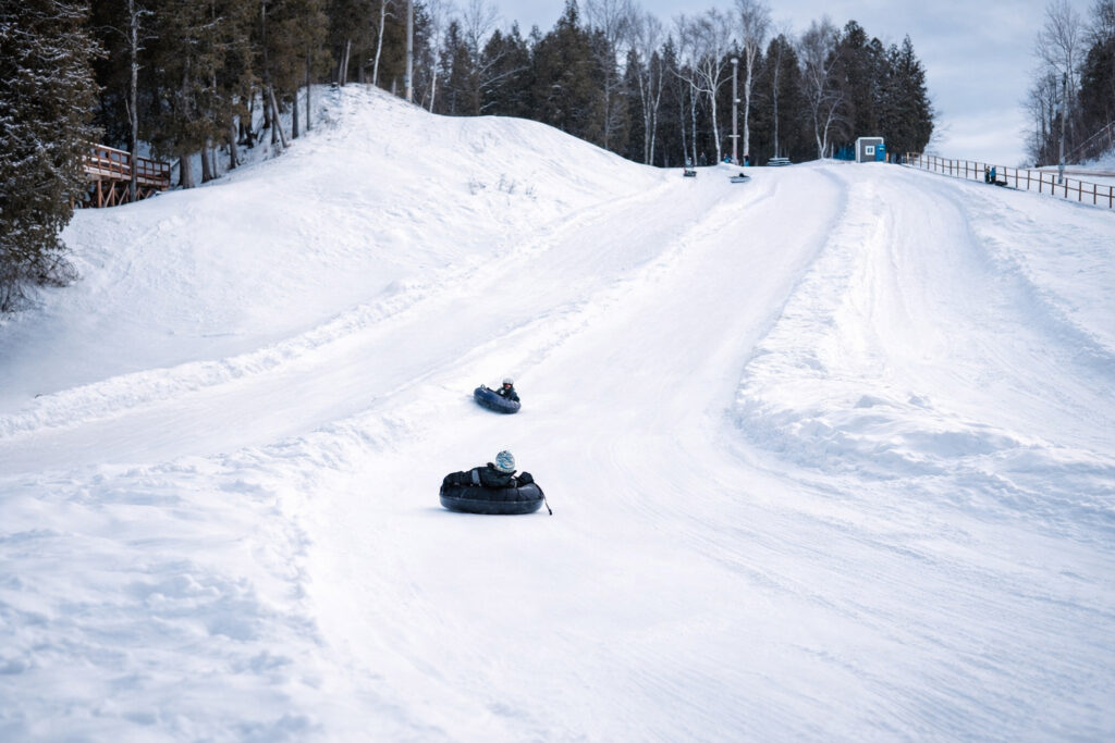 Three people are snow tubing down a wide, snowy hill at the Lake Superior Ice Festival, surrounded by trees on a winter day. The overcast sky and hill, bordered by fences and a blue hut at the top, create a festive winter scene.