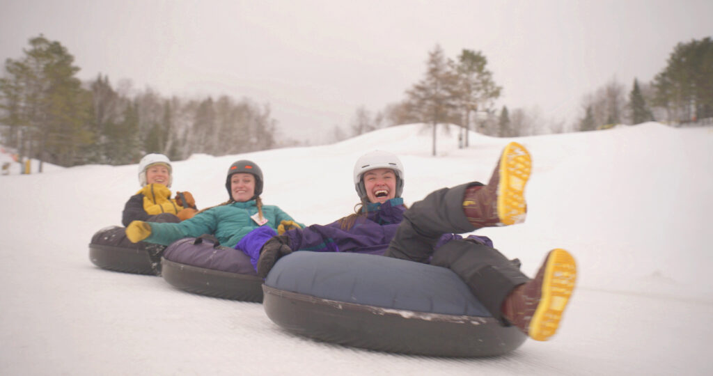 Three people wearing winter gear and helmets are laughing and sliding down a snowy hill on large inner tubes at the Lake Superior Ice Festival, holding onto each other in a row. Snow-covered trees and a cloudy sky are in the background.