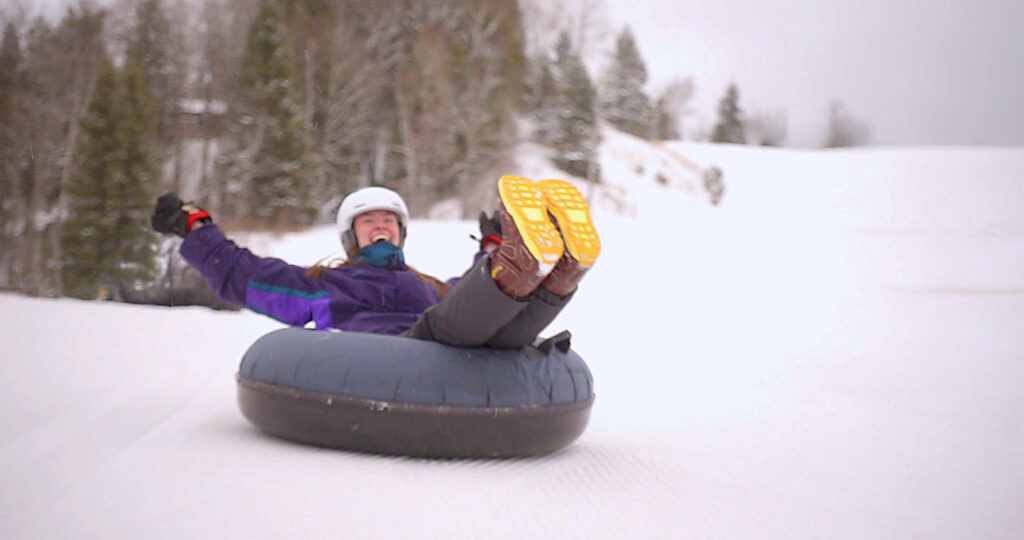 A person wearing a helmet and winter clothes slides down a snowy hill on an inflatable snow tube at the Lake Superior Ice Festival, smiling with arms raised and legs in the air; evergreen trees stand in the background.