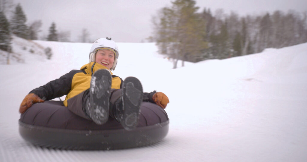A person wearing a yellow jacket, black pants, gloves, and a white helmet is smiling while sliding down a snowy hill on a black inflatable snow tube at the Lake Superior Ice Festival. Snow-covered trees are visible in the background.