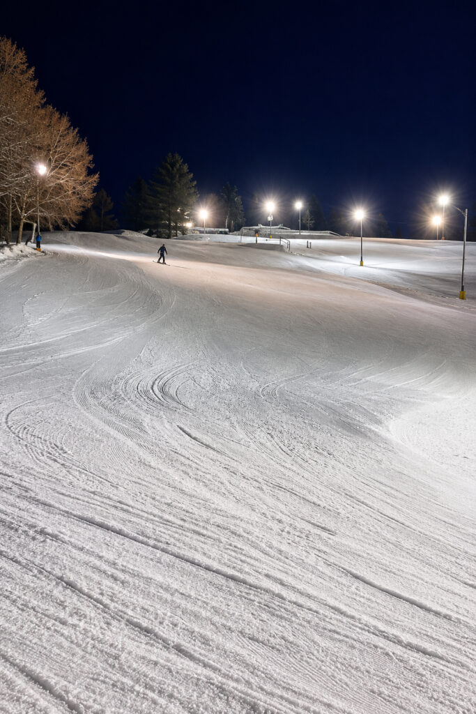 A skier descends a wide, snow-covered slope at night under bright floodlights during the Lake Superior Ice Festival, with tracks visible in the snow and trees lining the left side of the hill.