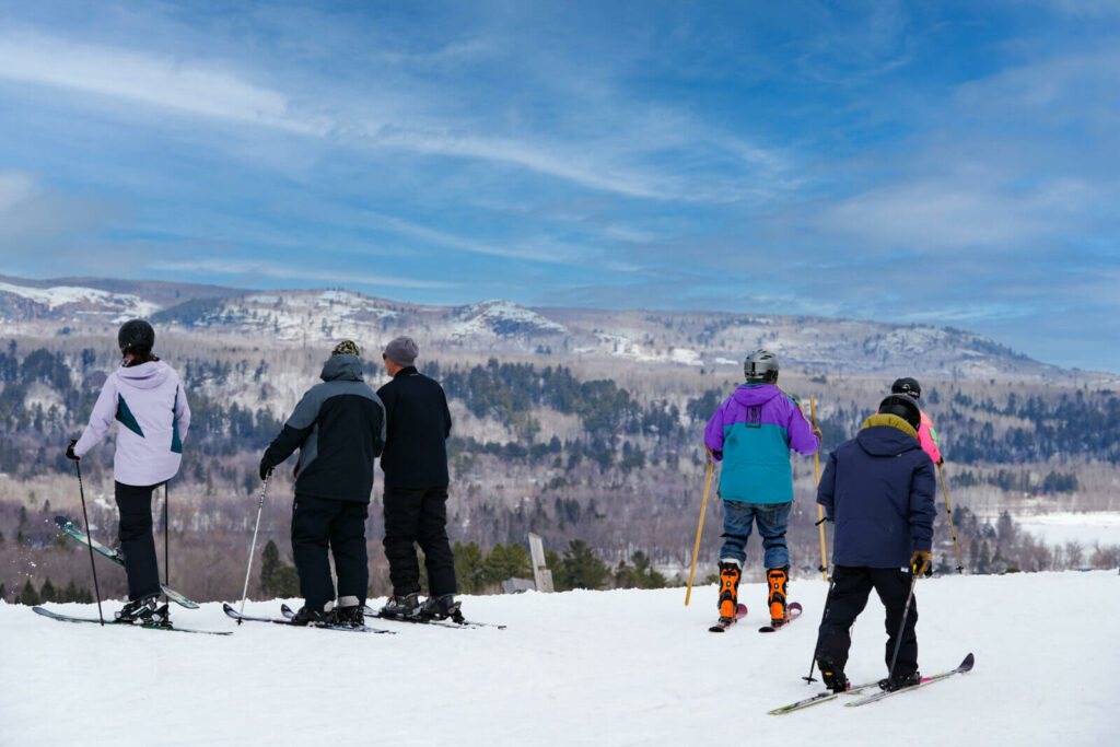 Six skiers in winter gear stand on a snowy slope, facing away and looking toward distant snow-covered mountains beneath a partly cloudy blue sky, capturing the adventurous spirit of the Lake Superior Ice Festival.
