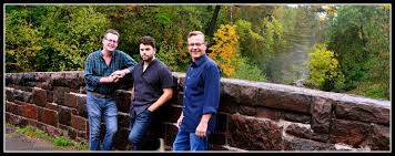 Three men stand and lean against a stone bridge railing, smiling. Behind them is a lush forest with trees in shades of green and yellow, suggesting early autumn. The mood is relaxed and casual.