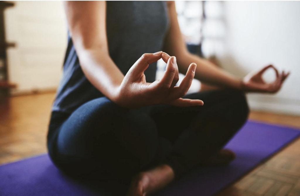 A person sitting cross-legged on a purple yoga mat indoors, wearing dark workout clothes, with hands resting on knees in a meditation pose, fingers forming a mudra.