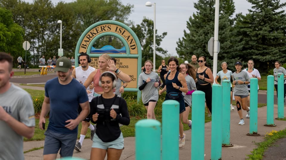 A group of people are jogging or running past a Barker's Island sign on a paved path surrounded by trees and teal posts, on a cloudy day.
