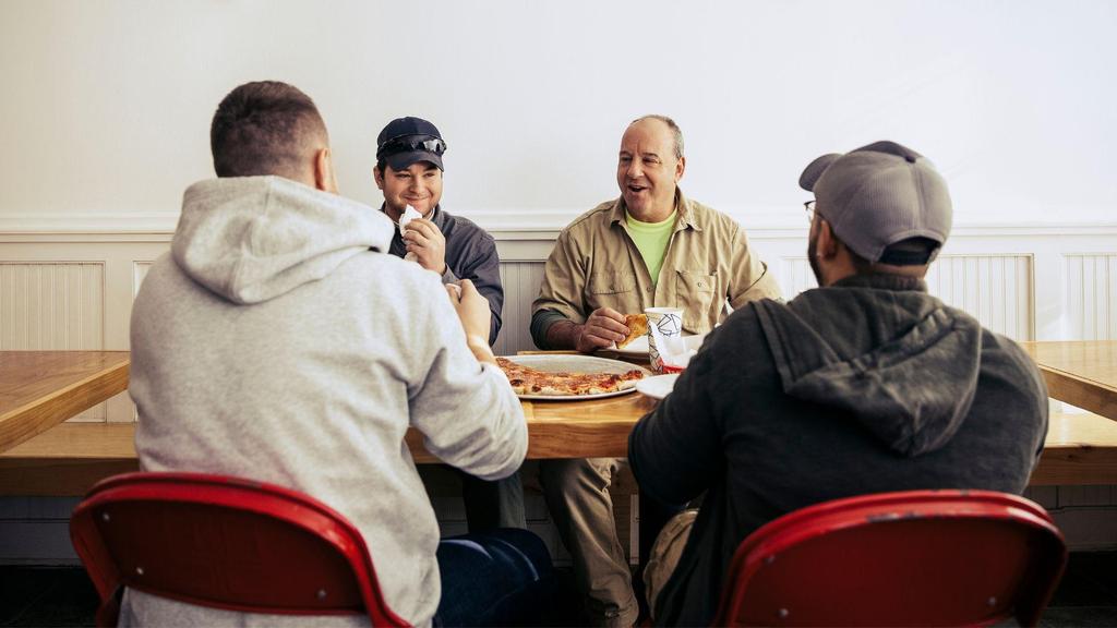 Four men sit around a wooden table with red chairs, sharing a pizza and drinks. They appear to be talking and laughing together in a casual, brightly lit setting.