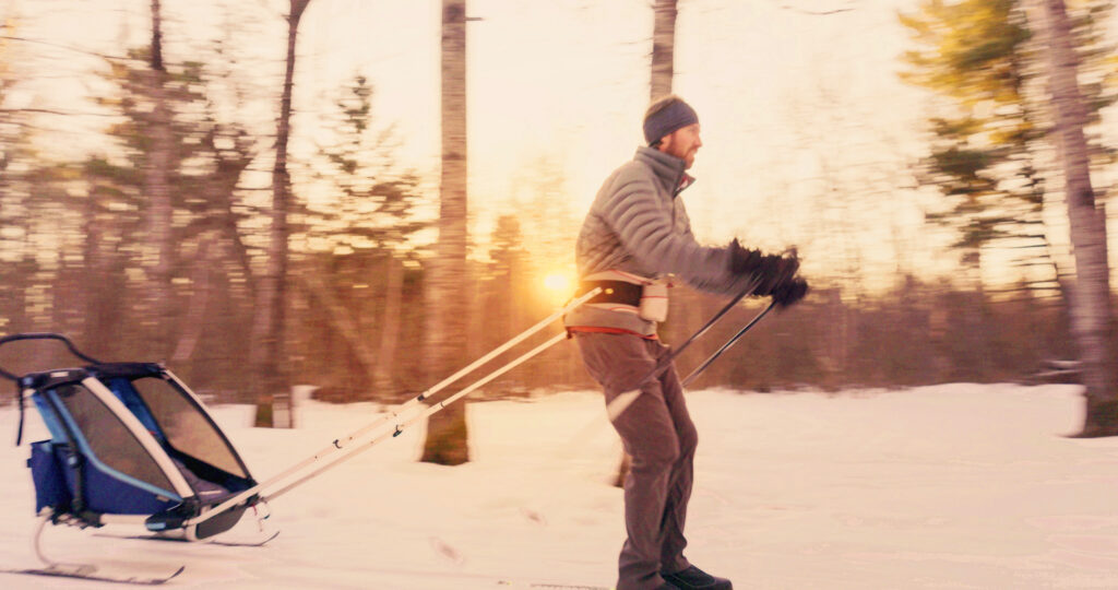 A person in winter clothing pulls a small sled through a snowy forest at sunset, with trees and sunlight in the background—capturing the spirit of adventure found at events like the Lake Superior Ice Festival.