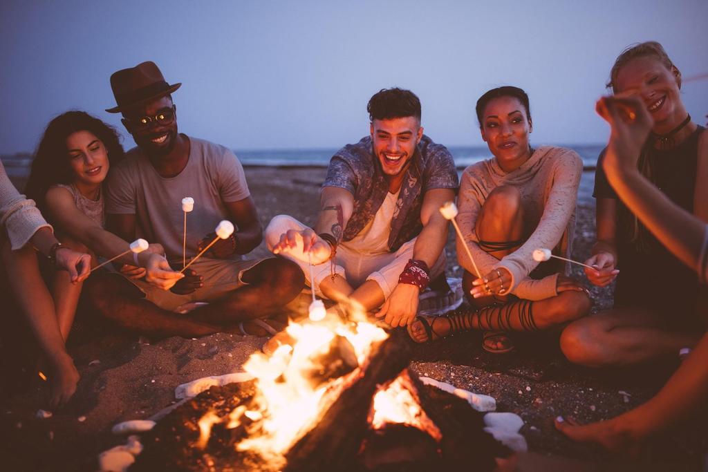 A group of friends sit in a circle on the beach at dusk, roasting marshmallows over a campfire. They are smiling and laughing, enjoying the warm fire and each other’s company. The ocean is visible in the background.
