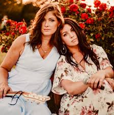 Two women sit closely together outdoors in front of red roses. One, in a light blue dress, holds a ukulele, while the other, in a floral dress, holds a microphone and leans her head on her companion’s shoulder.