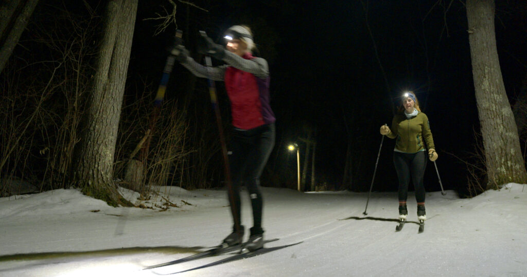 Two people cross-country ski at night on a snowy forest trail during the Lake Superior Ice Festival, wearing headlamps and winter clothing. Trees line the path, and a lamp post lights part of the background.