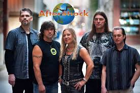 Five members of the band Aftershock stand together outdoors, posing for a group photo. The band’s logo with a globe is above them. They are casually dressed, with the woman in the center. The background is blurred.