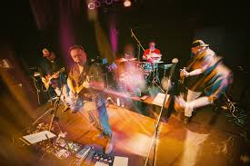 A band performs on stage under colorful lights. Four musicians play guitar, bass, and drums, with vibrant motion blur capturing the energy and movement of the live performance.