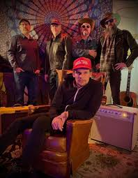 Five men pose in a room with musical instruments and patterned wall art; four stand in the background, while one sits in a brown armchair in front, wearing a red cap and black jacket.