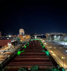 A nighttime view of an industrial area with illuminated buildings, large tanks, and green lights outlining pathways; the sky is dark and city lights are visible in the distance.
