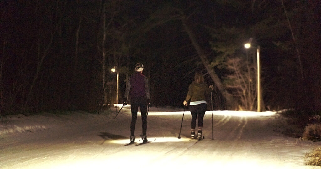 Two people are cross-country skiing at night on a snow-covered trail illuminated by several streetlights, surrounded by trees, enjoying the wintry magic of the Lake Superior Ice Festival.