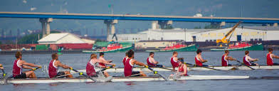 Two teams of rowers in red uniforms compete in a rowing race on a river, with industrial buildings, a bridge, and boats visible in the background.
