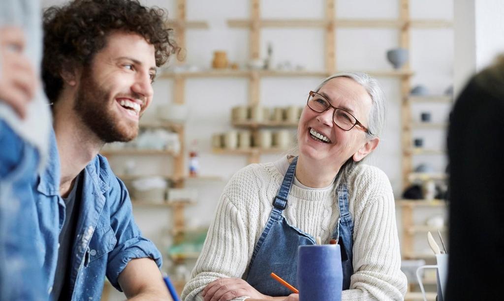 Two people, one older woman in glasses and an apron and one younger man with curly hair and a beard, are smiling and talking together at a table in a pottery or art studio. Shelves with pottery are in the background.