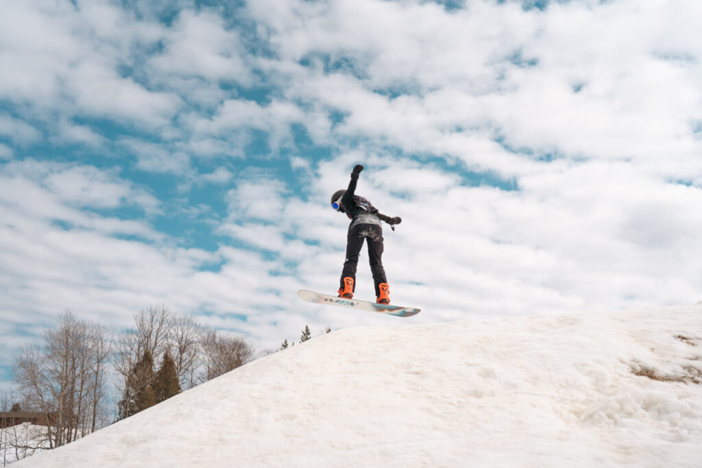 A snowboarder in mid-air performing a jump on a snowy slope at the Lake Superior Ice Festival, with trees in the background and a blue sky filled with scattered clouds overhead.