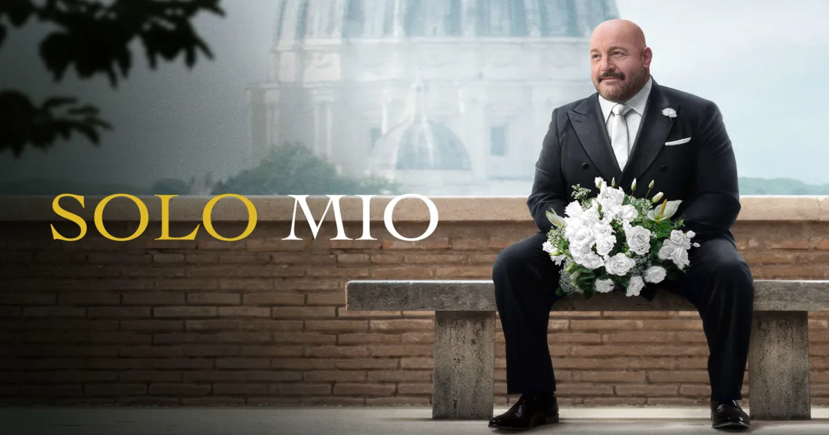 A bald man in a black suit sits on a stone bench holding a bouquet of white flowers. Behind him is a blurred domed building. The words "SOLO MIO" appear to the left of him.