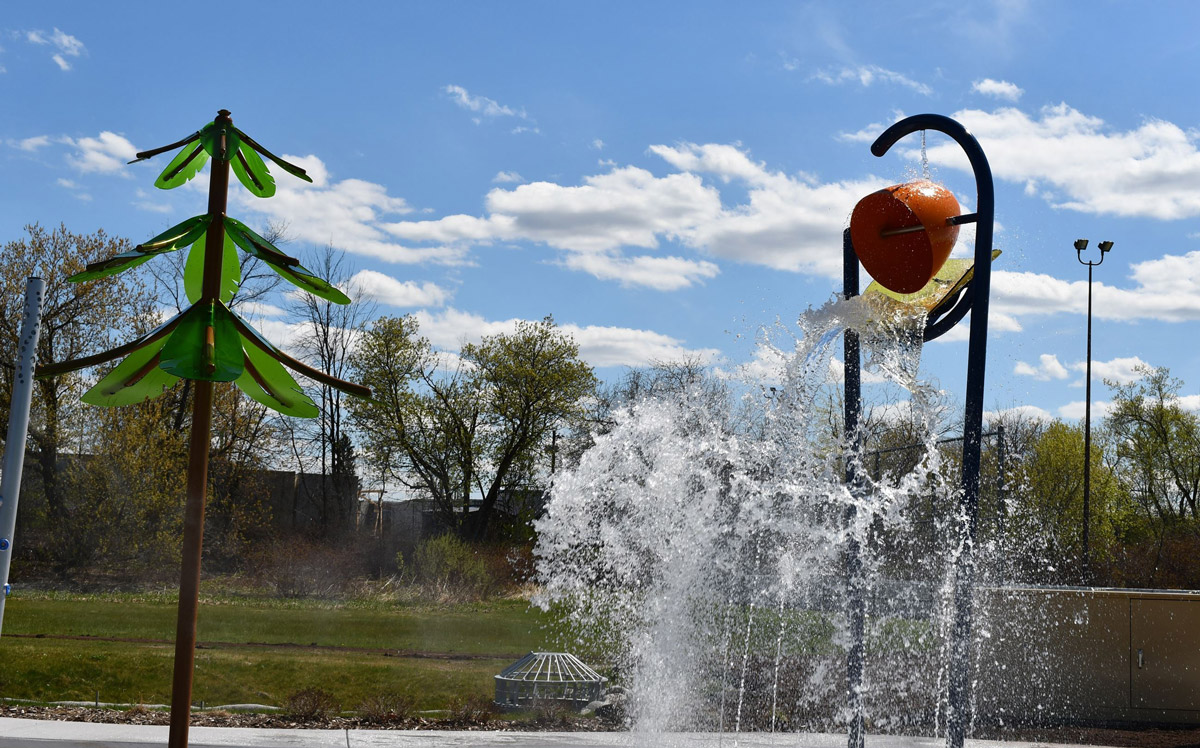 A large orange bucket tips over, spilling water onto a splash pad under a blue sky with scattered clouds. A green palm tree-shaped structure stands nearby, with trees and grass in the background.