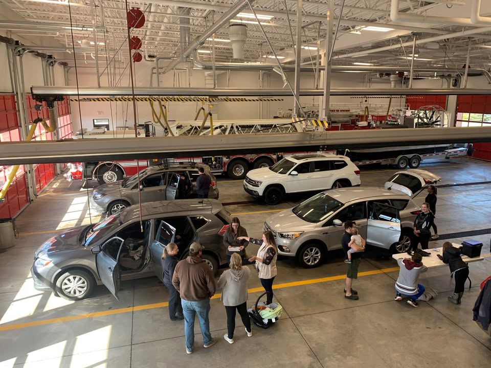 People are gathered around several cars inside a brightly lit garage, some cleaning or inspecting the vehicles. The garage has large windows, red doors, and fire equipment visible in the background.