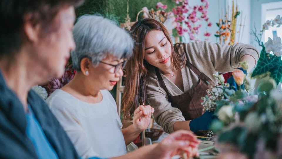 Three women working together on a flower arrangement in a bright room filled with plants and flowers. One woman arranges flowers while the others observe and assist.