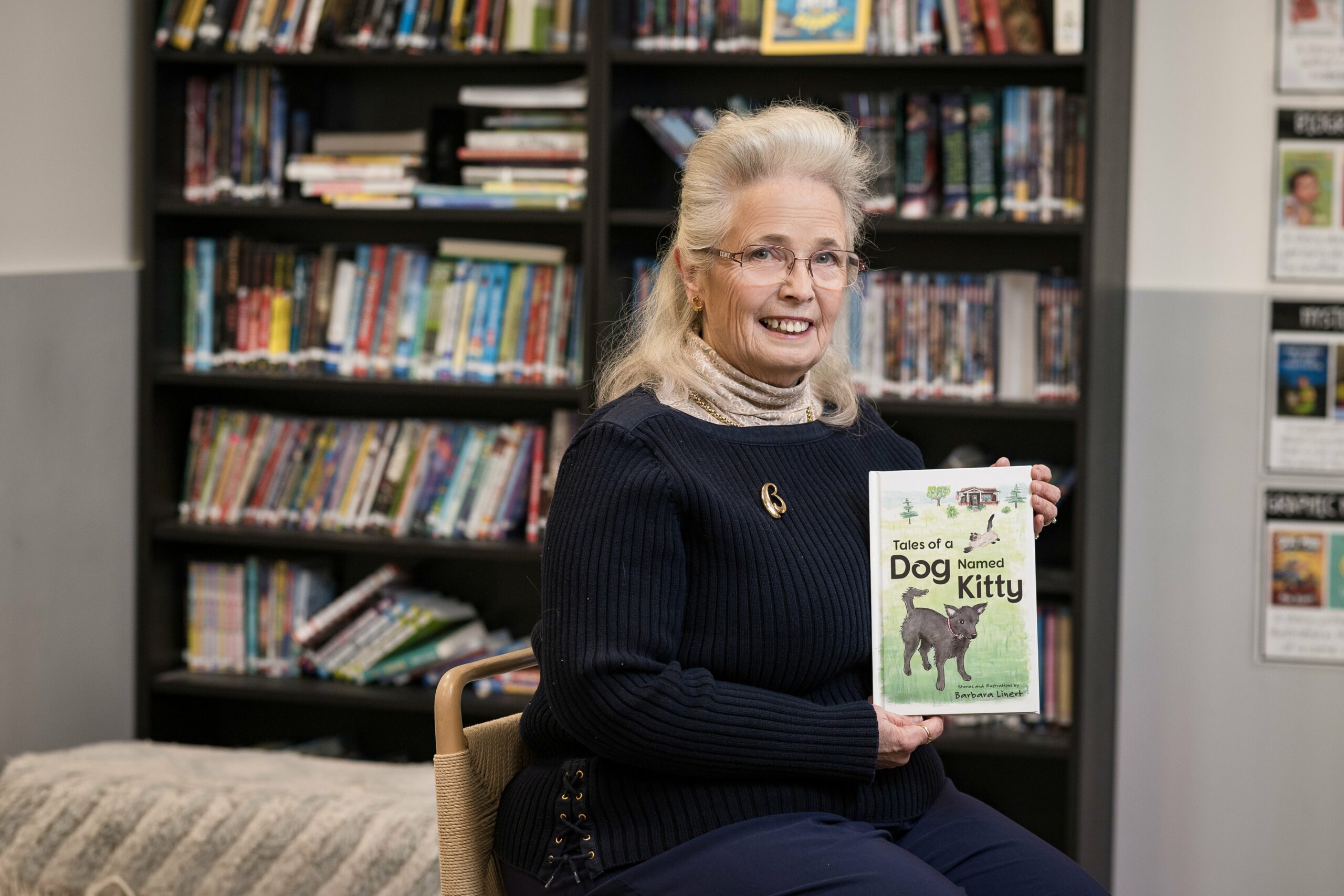 A woman with blonde hair and glasses sits in a library, smiling and holding a book titled “Tales of a Dog Named Kitty.” Shelves of books are visible in the background.