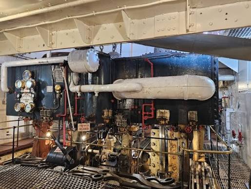 Large industrial steam engine with metal pipes, gauges, and valves in an enclosed room. Grated floor and safety railings visible, with machinery painted in dark and light colors. Overhead lighting illuminates the scene.