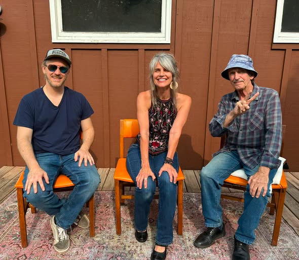 Three people sit on orange chairs outside, posing and smiling. The man on the left wears sunglasses and a cap, the woman in the middle has long gray hair, and the man on the right wears a bucket hat and flashes a peace sign.