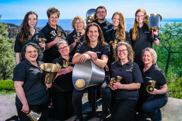 A group of eleven people, dressed in black, smile while holding handbells outdoors with a scenic, leafy background and blue sky.